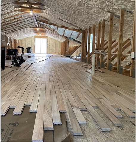 Hardwood planks being laid across a long attic room under insulated rafters