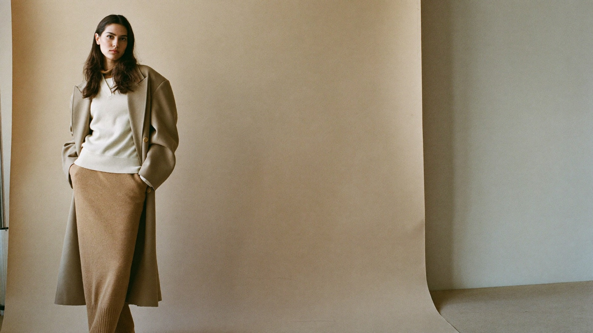 STRATA model in tonally layered knitwear and a tailored overcoat against a bone studio backdrop