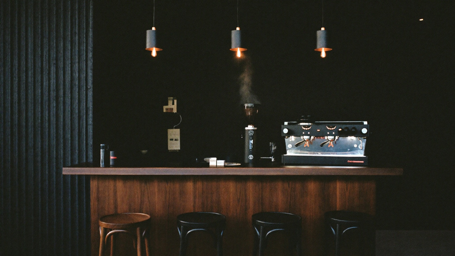 The dimly lit espresso bar at Verso Coffee Roasters — copper pendant lights over a walnut counter and a matte black espresso machine